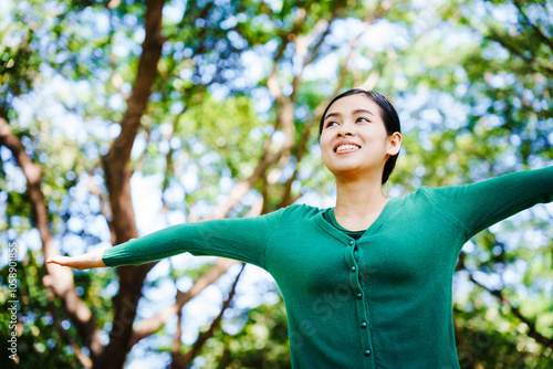 Young Asian woman sits under a shady tree in a park, enjoying her free time in nature. She breathes deeply, refreshing herself, surrounded by leaves and the tranquility of greenery.