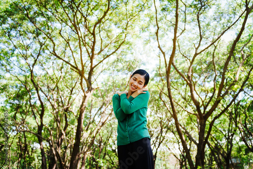 Young Asian woman sits under a shady tree in a park, enjoying her free time in nature. She breathes deeply, refreshing herself, surrounded by leaves and the tranquility of greenery.