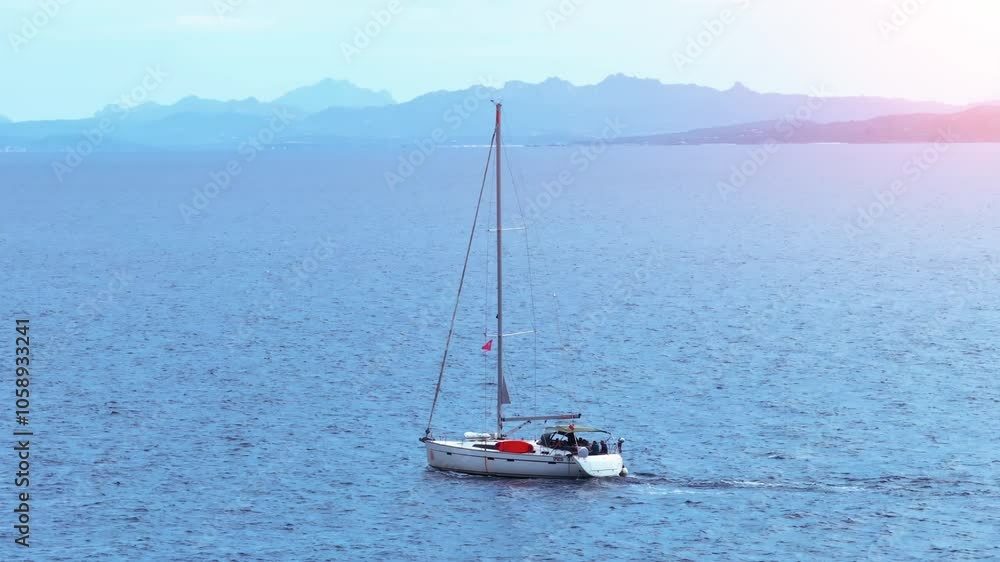 Sailboat cruising calmly on a tranquil ocean near distant mountains in the late afternoon sunlight with clear blue skies above