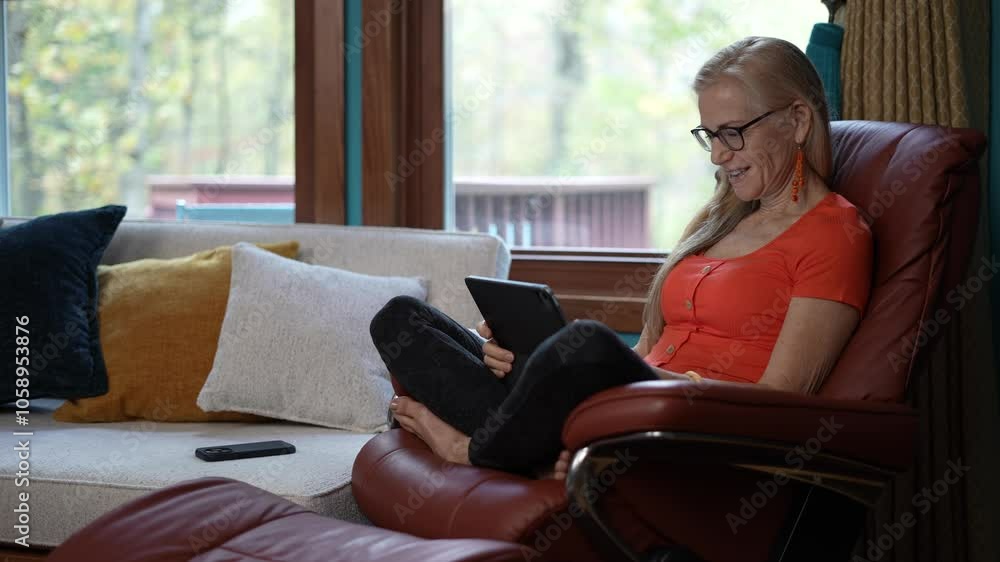 Surprised woman with long blonde hair, wearing glasses and an orange shirt, sits in a brown leather recliner with a tablet in her lap. She is looking at the tablet with an expression of surprise. Surprised woman with long blonde hair, wearing glasses and an orange shirt, sits in a brown leather recliner with a tablet in her lap. She is looking at the tablet with an expression of surprise.