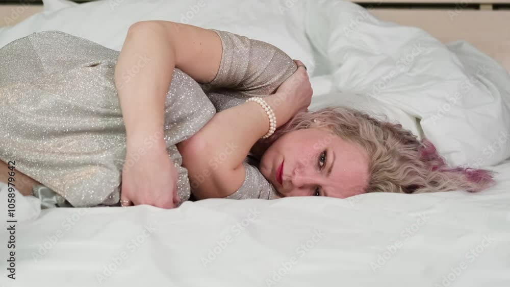 A woman with bright multi-colored hair in a beautiful shiny plaite is lying in bed after a party, very sad and disappointed. Perhaps she feels bad from drinking too much alcohol. A woman with bright multi-colored hair in a beautiful shiny plaite is lying in bed after a party, very sad and disappointed. Perhaps she feels bad from drinking too much alcohol.