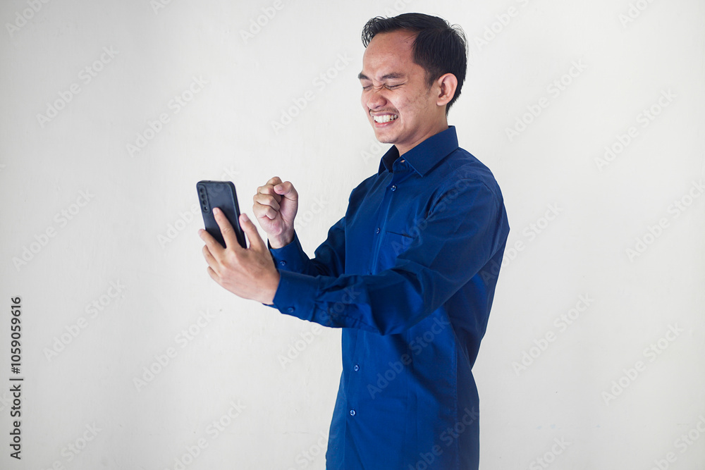 Smiling young Asian businessman in a blue shirt with a happy, surprised and happy expression while holding his smart phone on a white background. Ideal for technology, communication, social media enga