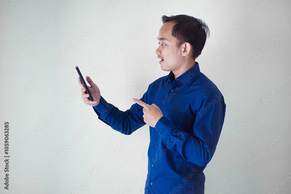 Smiling young Asian businessman in a blue shirt with a happy, surprised and happy expression while holding his smart phone on a white background. Ideal for technology, communication, social media enga Smiling young Asian businessman in a blue shirt with a happy, surprised and happy expression while holding his smart phone on a white background. Ideal for technology, communication, social media enga