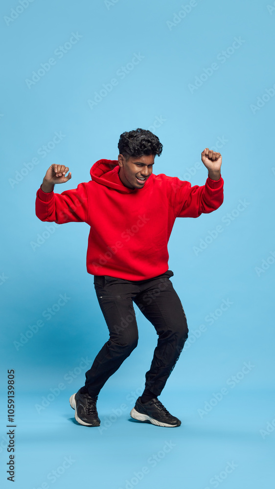 Full-length portrait of cheerful young Indian man in black jeans and red hoodie, cheerful dancing against blue studio background. Positive vibes. Concept of emotions, lifestyle, party, celebration Full-length portrait of cheerful young Indian man in black jeans and red hoodie, cheerful dancing against blue studio background. Positive vibes. Concept of emotions, lifestyle, party, celebration