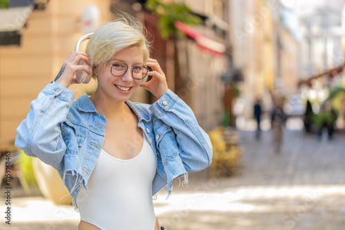 Girl in glasses taking off wireless headphones looking at camera smiling on city street. Relaxed young woman with short blond hair listening favorite energetic disco rock n roll music dancing outdoors