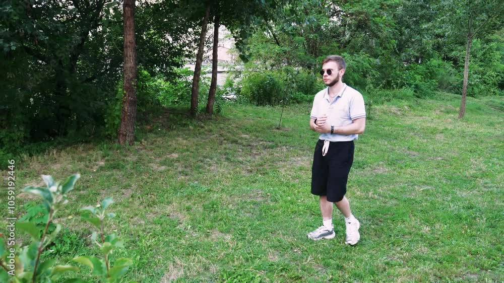A young man in sunglasses calmly strolls through a grassy area surrounded by plants and trees. Ideal for outdoor content. A young man in sunglasses calmly strolls through a grassy area surrounded by plants and trees. Ideal for outdoor content.