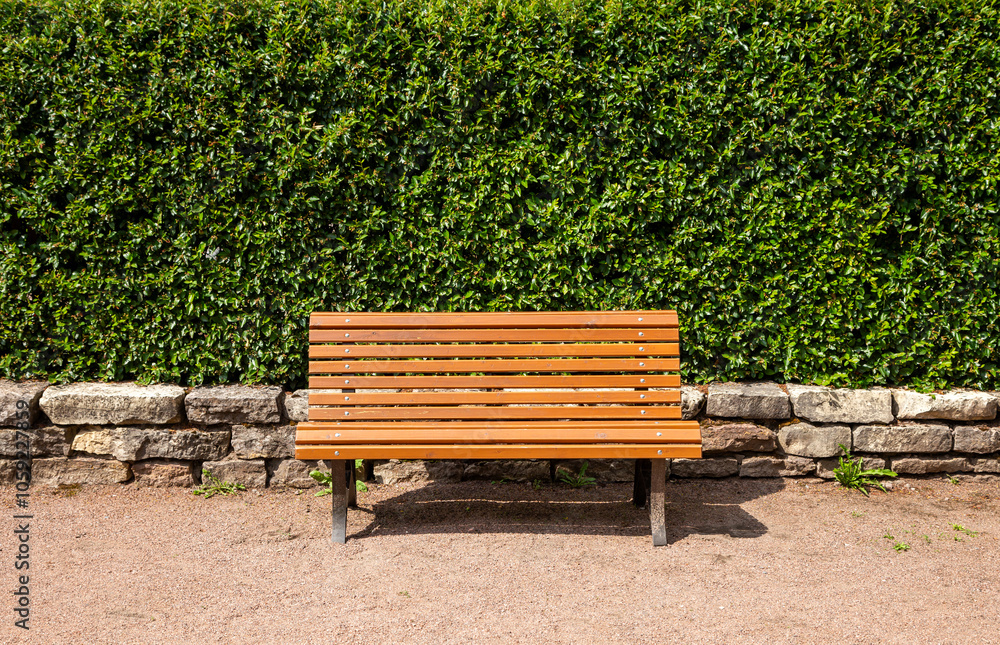 Brown wooden bench stands in a park against a hedge and low stone fence on a clear sunny day. Brown wooden bench stands in a park against a hedge and low stone fence on a clear sunny day.