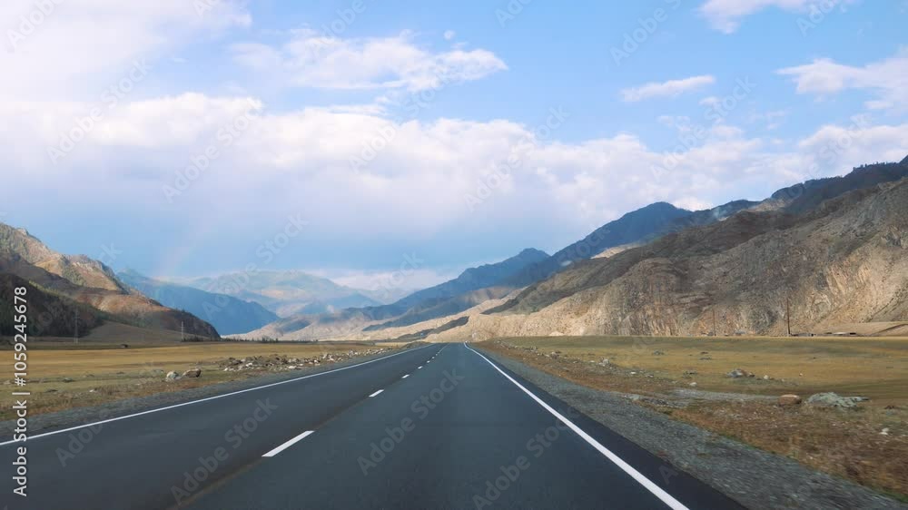 Driving on empty asphalt road winds through mountain valley, surrounded by yellow grass in prairie under sunny sky filled with clouds. View from vehicle driving along the highway. Road trip in summer Driving on empty asphalt road winds through mountain valley, surrounded by yellow grass in prairie under sunny sky filled with clouds. View from vehicle driving along the highway. Road trip in summer