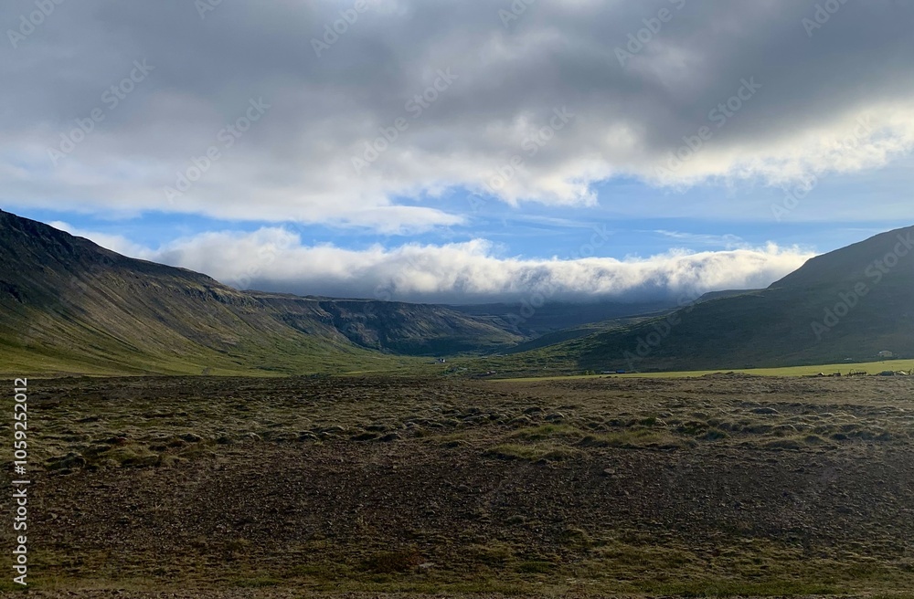 Fototapeta premium A ast valley with rugged mountains and low-hanging clouds, creating depth and isolation. Sunlight highlights the contours of the landscape, emphasizing Iceland's remote, serene beauty.