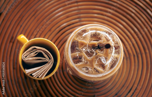 Top view of iced coffee in a plastic cup with a yellow mug containing paper napkin on a circular rattan table in a boho theme cafe