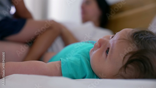 Close-up of a baby lying on a bed while the mother and young boy interact in the background, capturing a peaceful and relaxed family moment at home