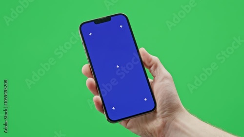 A modern smartphone is held in a man’s hand against a green background. The phone of the screen has a chromakey blue image with tracking markers.