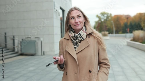 A woman walking outdoors in autumn attire while checking her phone near urban architecture during a sunny day