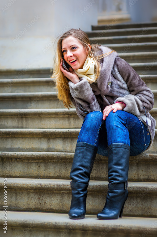 A woman in a fur-trimmed coat and jeans is seated on stone steps, smiling as she talks on her phone. Her posture is relaxed, and she seems engaged in a pleasant conversation..