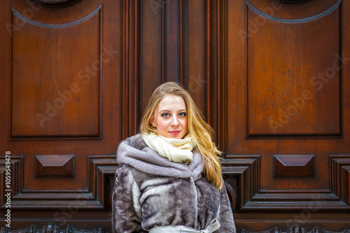 A woman with blonde hair and a warm, fur coat stands in front of an ornate wooden door. She wears a light-colored scarf and looks directly at the camera.