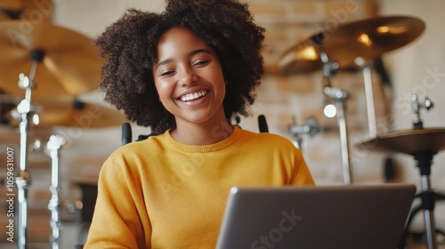 A woman with curly hair and a bright smile is sitting in a wheelchair, focused on working on her laptop in a music room with drums in the background. photo