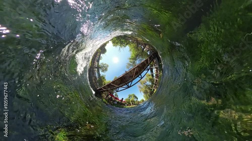 360 rotation. A fisheye view from beneath the wooden bridge, capturing the river's flow with sunlight reflecting on the water, surrounded by greenery and trees, creating a dynamic and immersive perspe photo