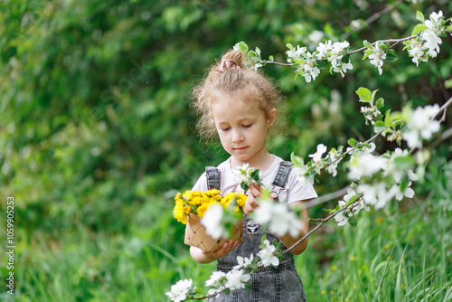 Little curly girl sniffing flowers of blossoming apple tree with dandellions in her hands. garden with flowering trees photo