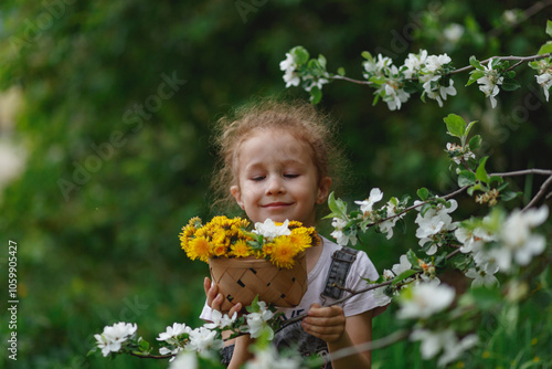 Little curly girl sniffing flowers of blossoming apple tree with dandellions in her hands. garden with flowering trees photo