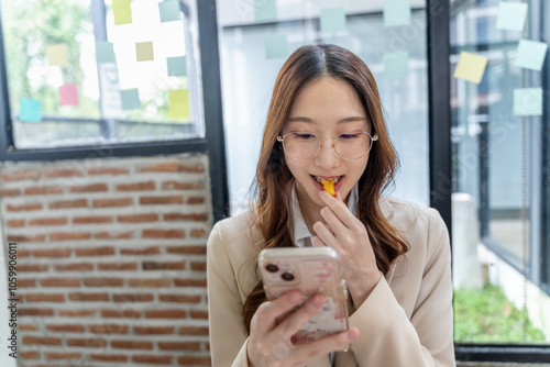 Happy young businesswoman eating french fried while using her phone at her office photo