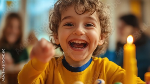 A young boy with curly hair laughs and points at the camera in a room with a lit candle in the background.