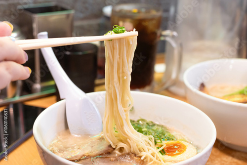 Hand holding chopsticks with yellow noodles of Hakata ramen with sliced pork, boiled egg and green spring onion in white bowl photo