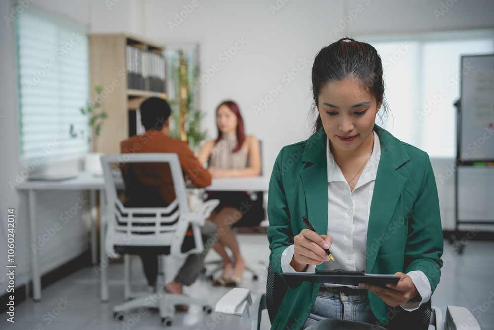Young asian businesswoman holding a digital tablet and taking notes while her colleagues are having a meeting in the background Young asian businesswoman holding a digital tablet and taking notes while her colleagues are having a meeting in the background