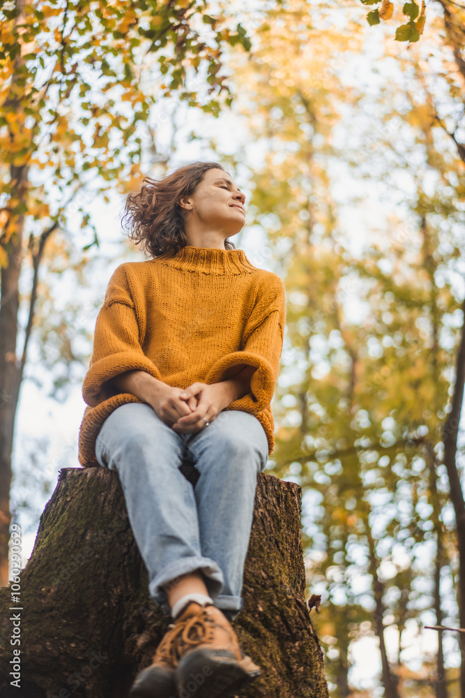 Young Caucasian woman enjoying autumn nature and weather sitting on a tree stump in autumn forest