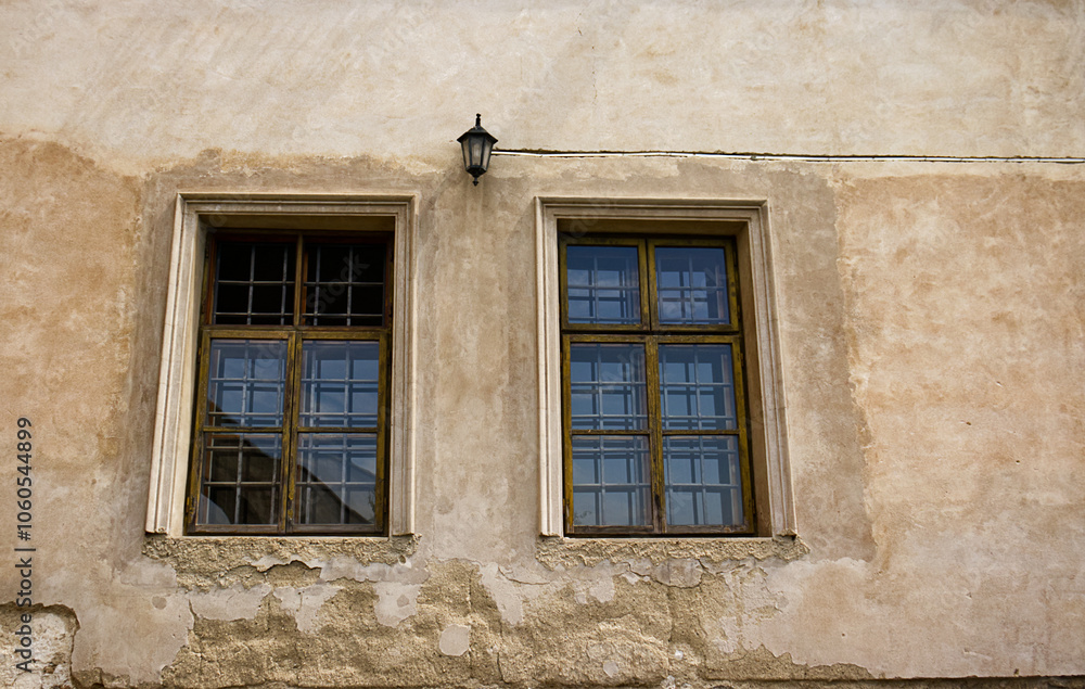 Two rectangular windows with bars. Windows of an old castle building. Decorative lamp on the wall above the windows. Old decaying facade. Windows, glass, lamp, fallen facade, rough, broken Two rectangular windows with bars. Windows of an old castle building. Decorative lamp on the wall above the windows. Old decaying facade. Windows, glass, lamp, fallen facade, rough, broken