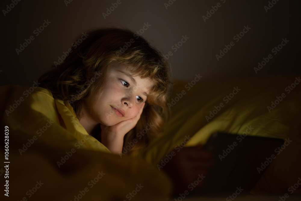 Kid is addicted to tablet, kid use tablet, watching cartoon. Child watching his tablet in the bed. Illuminated child face from device screen. Boy under the covers hold a tablet. Night time. Kid is addicted to tablet, kid use tablet, watching cartoon. Child watching his tablet in the bed. Illuminated child face from device screen. Boy under the covers hold a tablet. Night time.