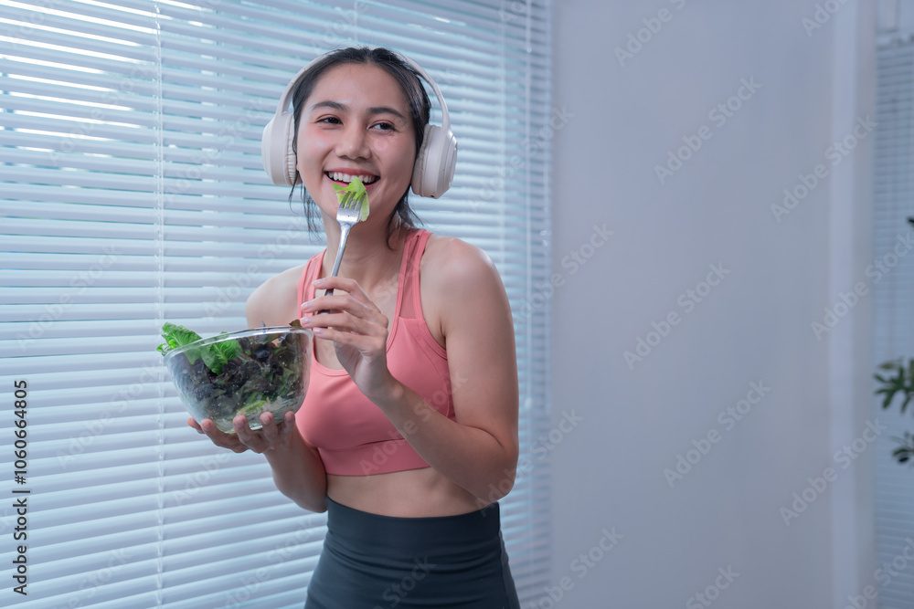 Smiling sporty asian woman wearing headphones enjoys a healthy salad while listening to music at home, promoting a balanced lifestyle combining fitness and nutrition Smiling sporty asian woman wearing headphones enjoys a healthy salad while listening to music at home, promoting a balanced lifestyle combining fitness and nutrition