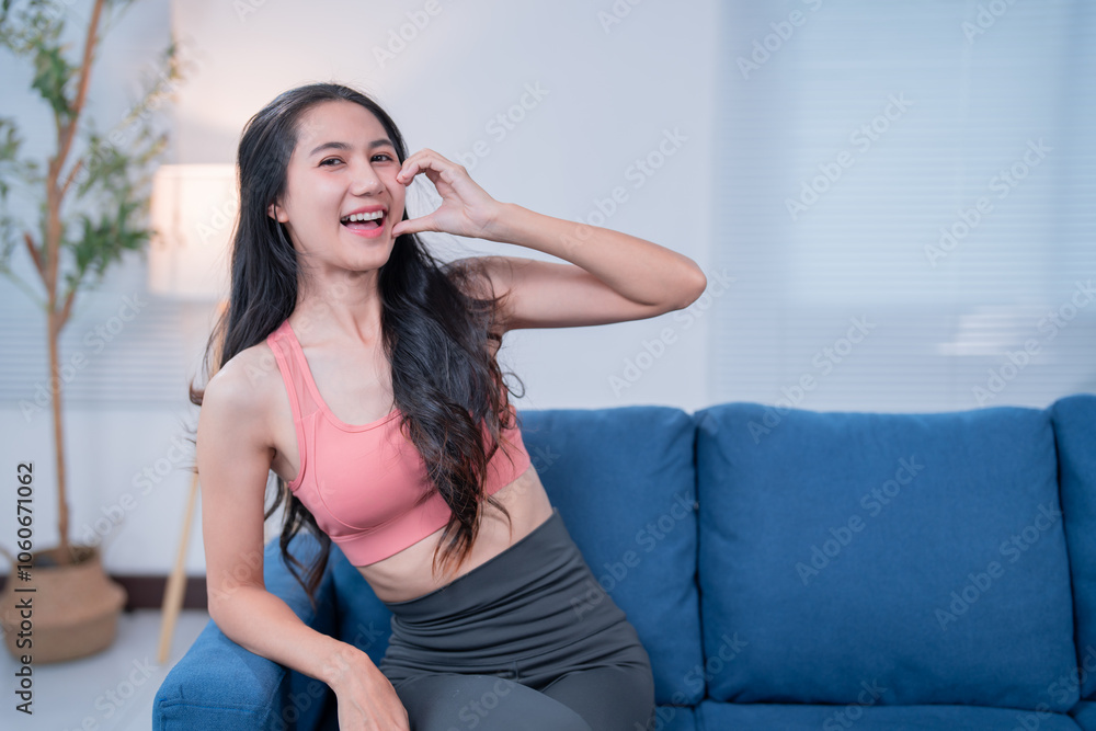 Happy athletic woman gesturing a phone call with her hand while sitting on a comfortable blue sofa in her modern apartment, enjoying a moment of relaxation and communication Happy athletic woman gesturing a phone call with her hand while sitting on a comfortable blue sofa in her modern apartment, enjoying a moment of relaxation and communication