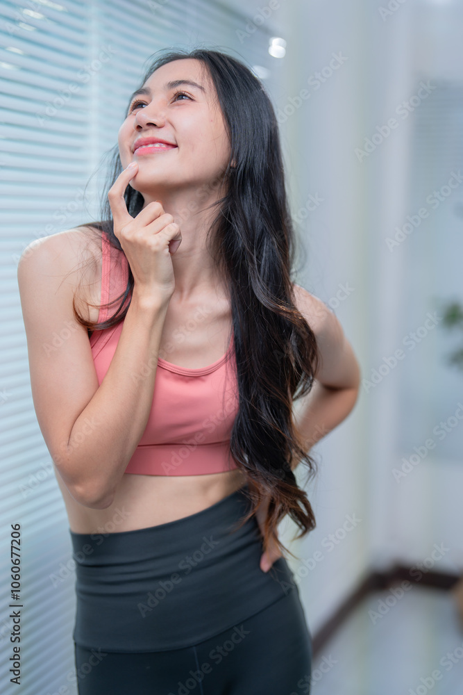 Asian woman in sportswear stands by a window at home, deep in thought, touching her chin with a happy smile, exuding wellness and contentment