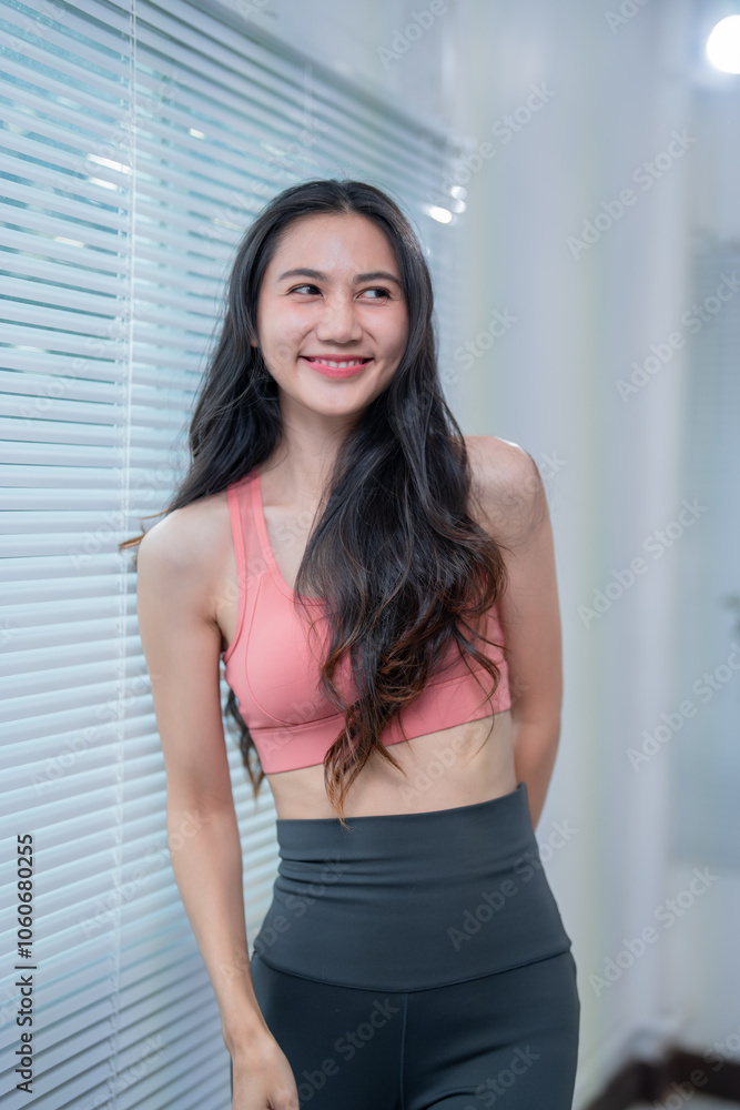 Happy sporty girl with long hair wearing sportswear standing next to window blinds, smiling and looking away after exercising at home or gym, enjoying healthy lifestyle and wellness Happy sporty girl with long hair wearing sportswear standing next to window blinds, smiling and looking away after exercising at home or gym, enjoying healthy lifestyle and wellness