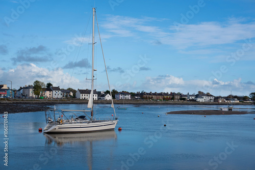 A serene coastal village harbor with a sailboat anchored in calm waters, under clear sky. Picturesque houses line shore, creating a peaceful idyllic scene. Not season for vacatio on a yacht