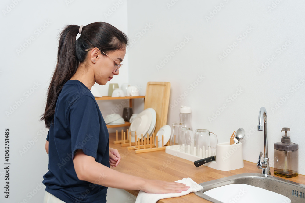 Young Asian woman drying kitchen countertop with cloth in neat, organized kitchen space, showing relaxed and focused expression, embodying calm, mindful homekeeping during personal time. Young Asian woman drying kitchen countertop with cloth in neat, organized kitchen space, showing relaxed and focused expression, embodying calm, mindful homekeeping during personal time.