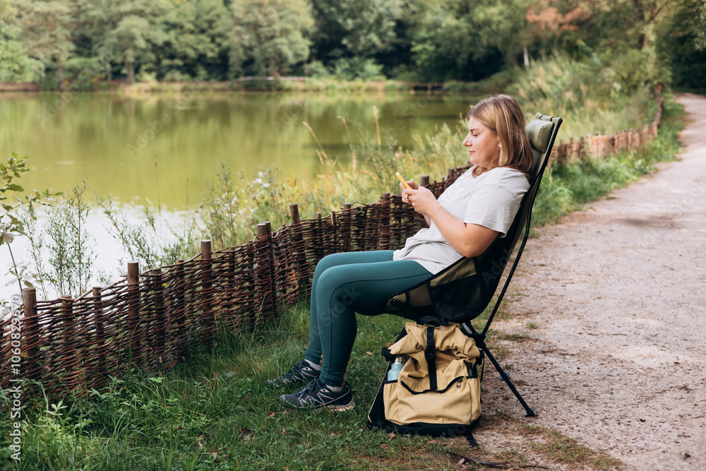 Relaxed happy woman sitting on deck chair near lake and holding mobile phone, she is planning new travel. Camping, tourism, technology and travel concept. A young 30s woman explores a forest and lake Relaxed happy woman sitting on deck chair near lake and holding mobile phone, she is planning new travel. Camping, tourism, technology and travel concept. A young 30s woman explores a forest and lake