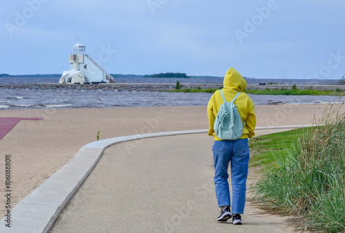 Lighthouse on a sandy beach in Oulu, Finland, on a cold, stormy spring day. In the foreground, a lonely girl with a blue backpack in a yellow sweatshirt walks on the beach, seen from behind. photo