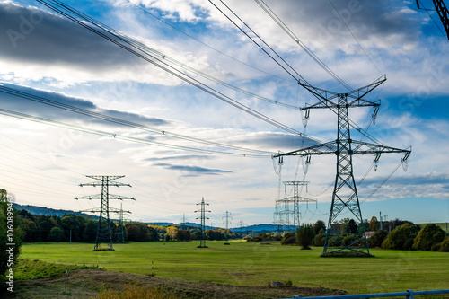 A landscape photo of power lines and transmission towers stretching across an open field with a scenic backdrop of hills and a partly cloudy sky. 