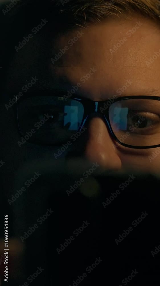 In a dimly lit room, an individual with glasses focuses intently on their smartphone, illuminated by its screen. The ambiance suggests a late-night setting In a dimly lit room, an individual with glasses focuses intently on their smartphone, illuminated by its screen. The ambiance suggests a late-night setting