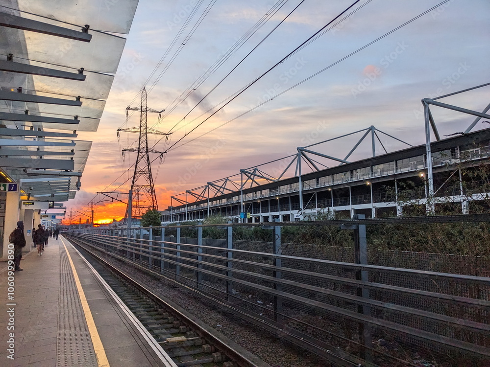 London - 03.14.2024: Passengers waiting for a train at the Custom House Elizabeth Line station behind the yellow line next to the Excel Exhibition Centre during sunrise with the sun at the horizon London - 03.14.2024: Passengers waiting for a train at the Custom House Elizabeth Line station behind the yellow line next to the Excel Exhibition Centre during sunrise with the sun at the horizon