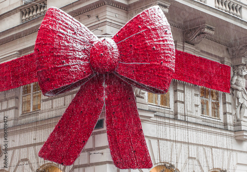 Canvas Print A huge red bow with a ribbon on the facade of a building in Christmas lights during a snowfall