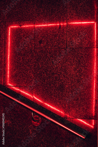 Red neon light frame is reflected on textured dark wall with a stair railing below, creating a bold contrast and modern look. Suitable for backgrounds, urban, or nightlife themes.
