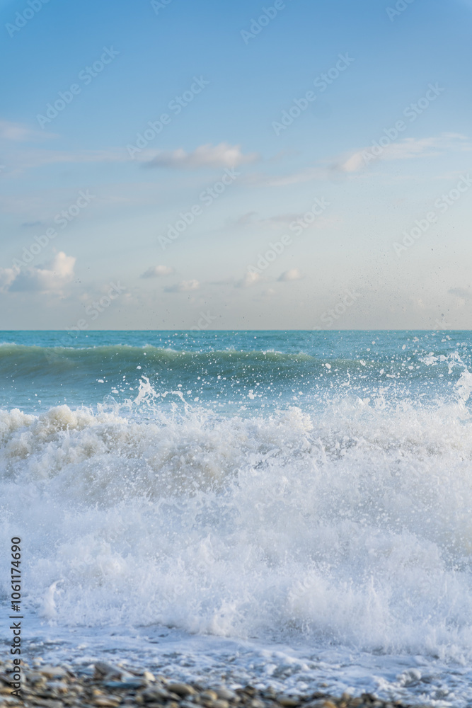 Fototapeta premium The sea waves roll onto a pebble sea shore against a blue sky with clouds.