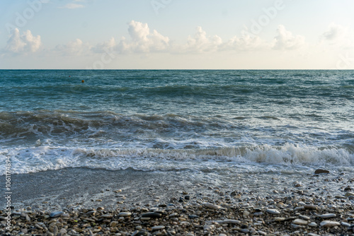 The sea waves roll onto a pebble sea shore against a blue sky with clouds.