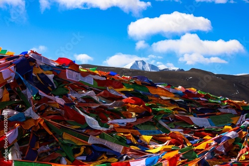 Vibrant Tibetan prayer flags flutter under a bright blue sky near the Himalayan snow-capped peaks at Pang La Pass, Tingri, Tibet, capturing the serene beauty of Tibetan spirituality