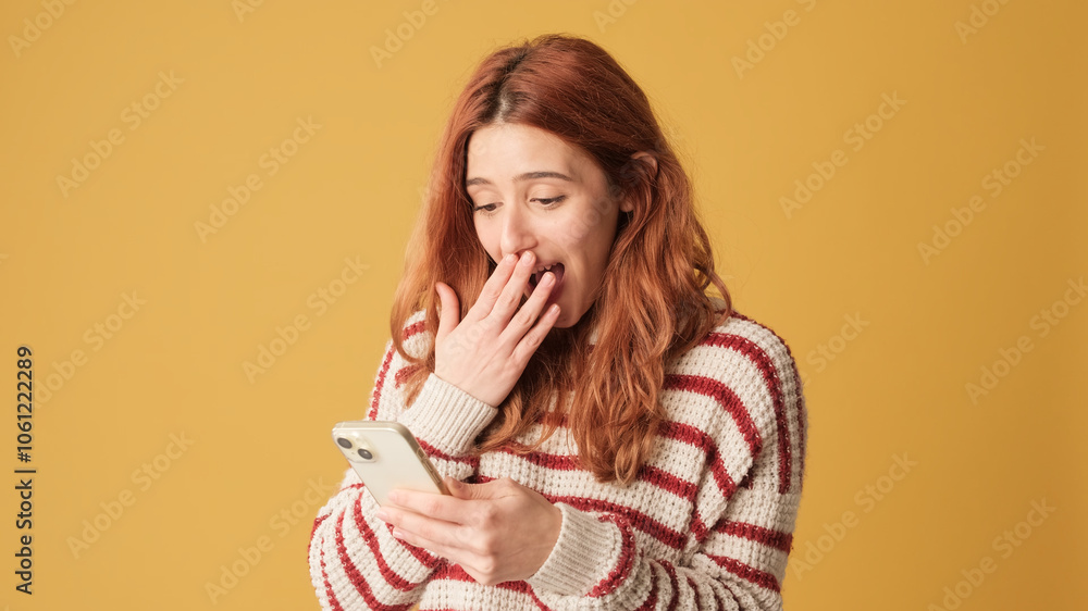 Surprised red-haired woman dressed in sweater, looking at camera while holding smartphone on yellow background. Beautiful girl poses in studio with blank space on yellow wall for your promotion Surprised red-haired woman dressed in sweater, looking at camera while holding smartphone on yellow background. Beautiful girl poses in studio with blank space on yellow wall for your promotion