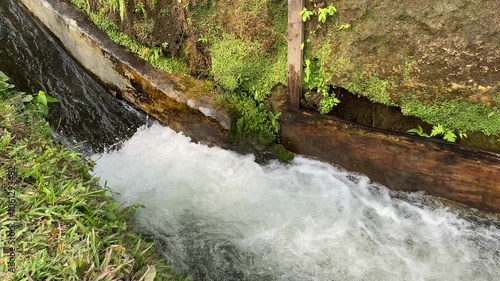 Close up view of river currents or water ripples. Clear fresh water flowing with wooden edge, green leaves plants, rocks and nature surrounding photo