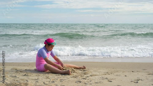 asian child summer at sea or kid girl happy fun sitting playing sand on beach with little wave for holiday travel or young people wearing swimsuit vacation relax at island in Thailand with wind sound