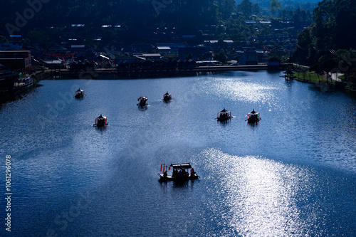 A traditional wooden boat adorned with red lanterns floats peacefully on the serene waters of Ban Rak Thai Village, surrounded by misty mountains at sunrise.	